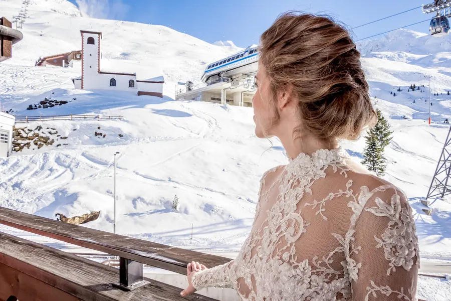 Braut im Spitzenkleid blickt auf verschneite Bergkapelle, Winterhochzeit am Arlberg